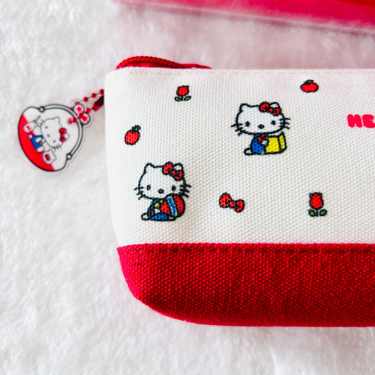 A close-up of a white canvas zip pouch with red trim and embroidered Hello Kitty characters, shown on a white textured background with a Hello Kitty keychain tag.