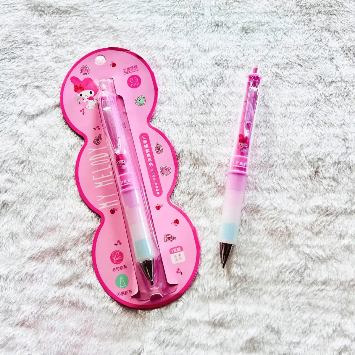 A pink and white My Melody mechanical pencil shown both in its pink blister packaging and unpackaged, resting on a white textured faux fur surface.