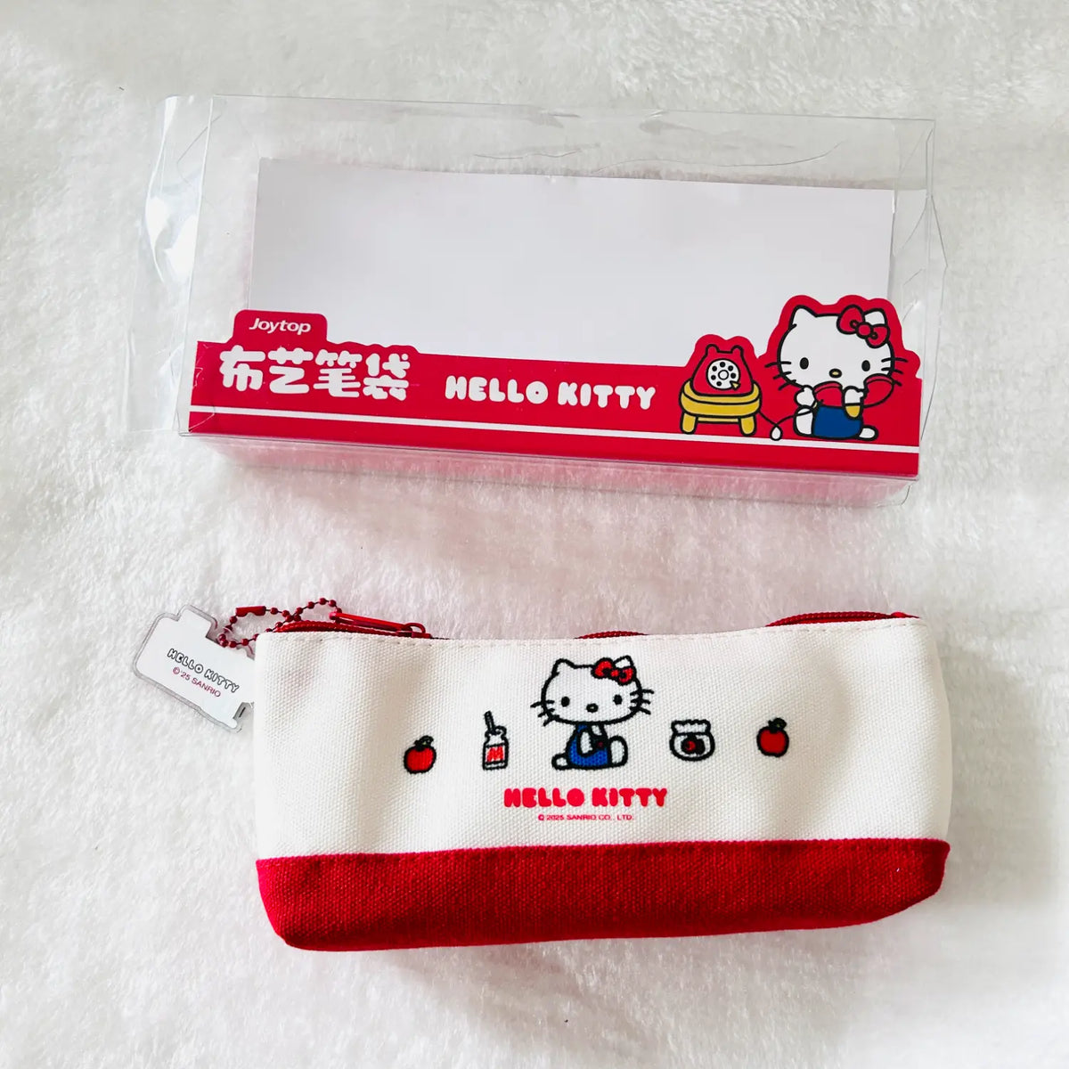 A Hello Kitty branded fabric pencil pouch shown below its clear plastic packaging box on a white textured background. The pouch is off-white with a red bottom band and features a printed Hello Kitty character holding a red apple, alongside small illustrations of an apple, a mug, and a pencil.