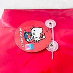 A red Hello Kitty branded circular sticker or seal attached to a red translucent plastic pouch with two clear plastic fastening clips on the side. The sticker features the Hello Kitty character and the text 'HELLO KITTY'.