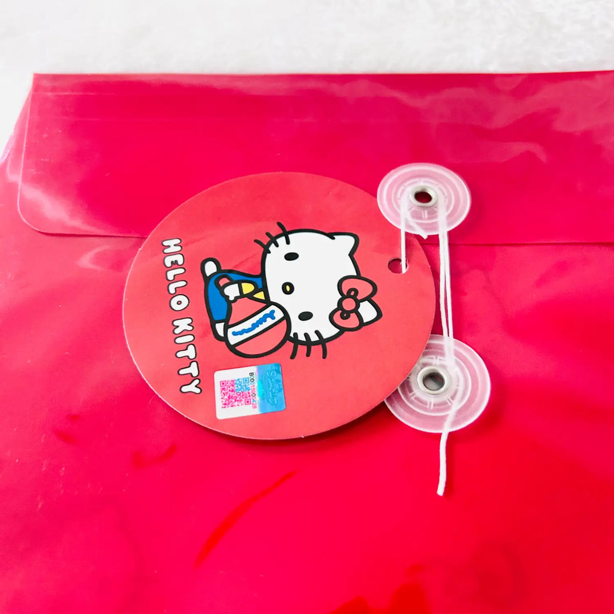 A red Hello Kitty branded circular sticker or seal attached to a red translucent plastic pouch with two clear plastic fastening clips on the side. The sticker features the Hello Kitty character and the text 'HELLO KITTY'.