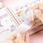 A close-up of hands peeling a transparent sticker sheet in front of a notebook. 