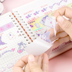 A close-up of hands peeling a transparent sticker sheet from a notebook. The sticker sheet features cute pastel illustrations, including a purple cartoon character and a playing card with hearts.