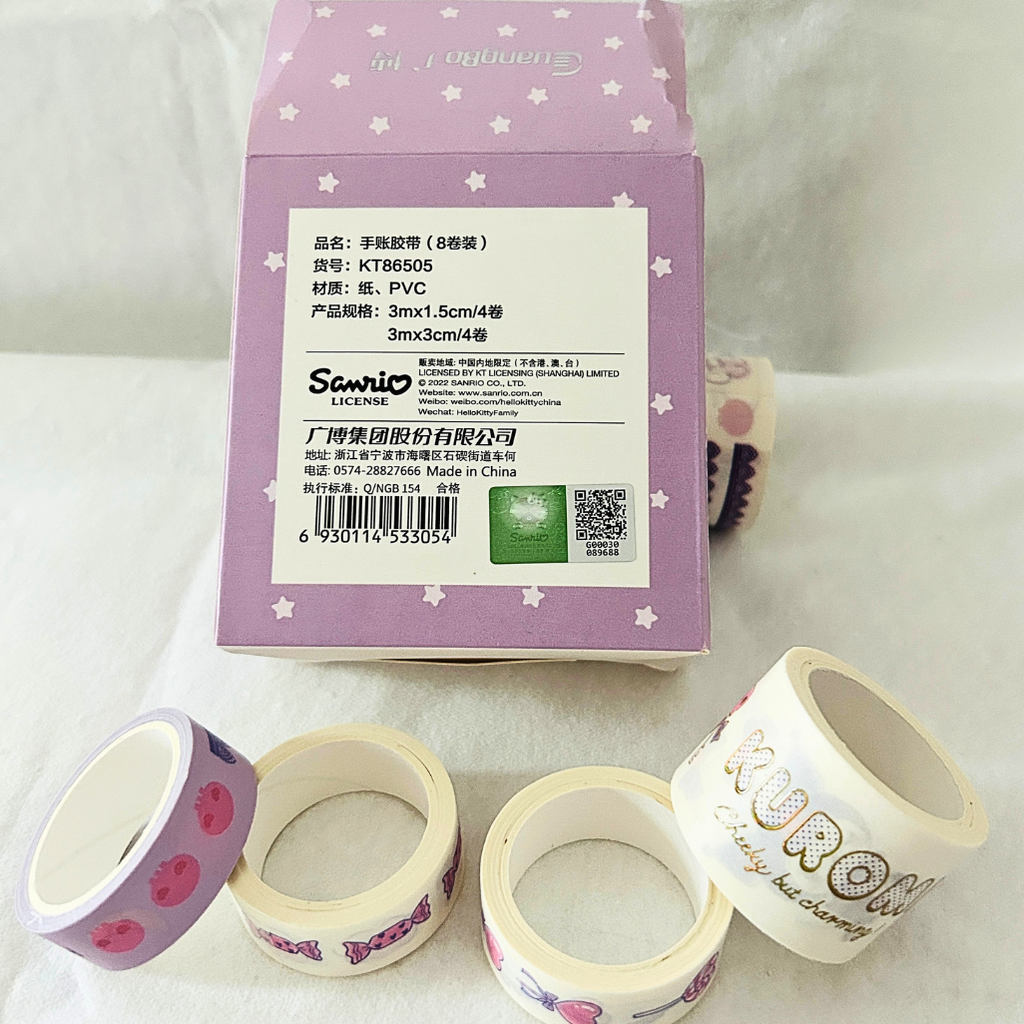 A box of Scenitio decorative washi tape with several rolls displayed in front on a white fabric background. The packaging is light purple with white stars, and the tape rolls feature pastel pink, cream, and white designs with floral and script patterns.