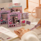 Kuromi-themed washi tape products displayed on a desk, including packaged rolls and loose rolls, with a person's hands holding a roll in front of an open notebook and a pink mug in the background.