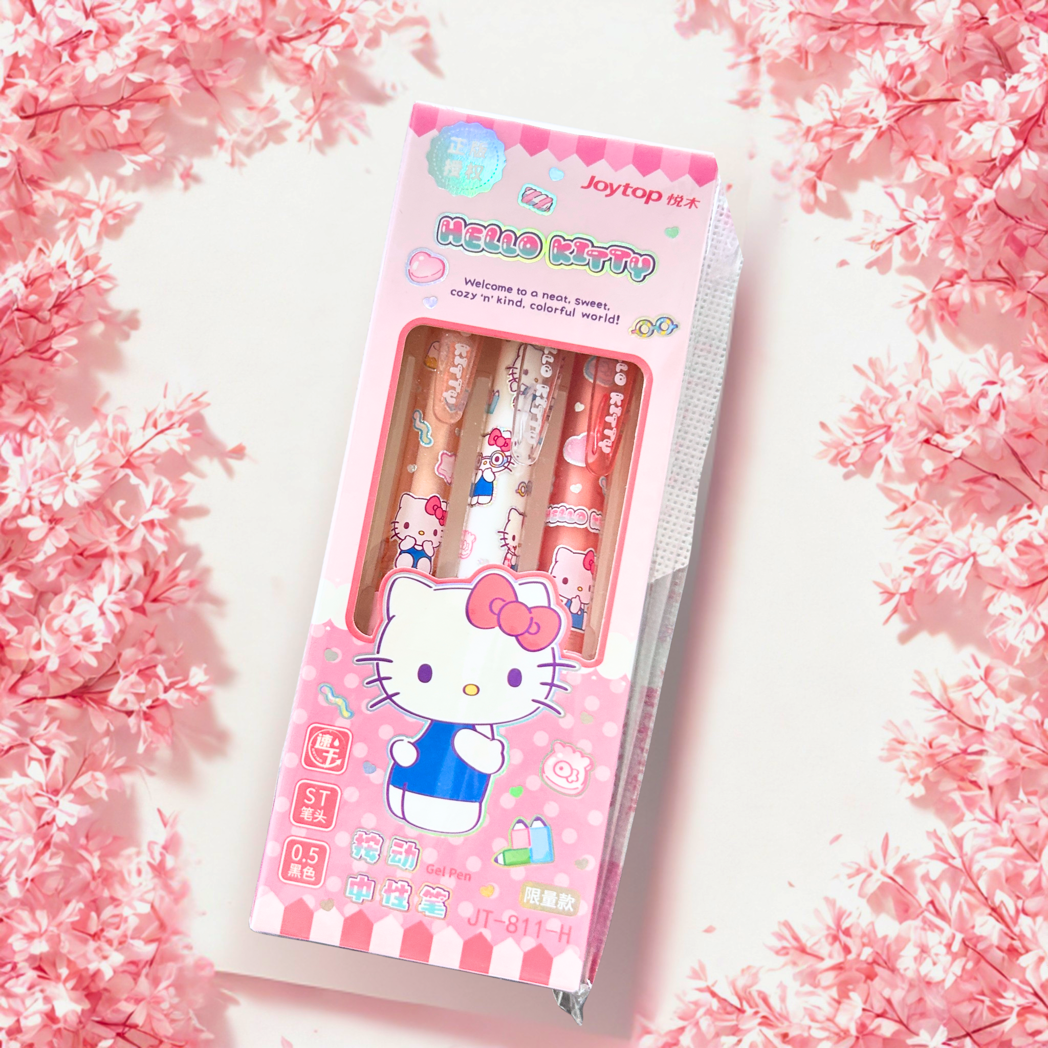 A boxed set of three Hello Kitty pens by Jaytop, displayed on a white surface surrounded by pink cherry blossoms. The packaging is predominantly pink with a large Hello Kitty illustration and a clear window showing the pastel-colored pens inside.
