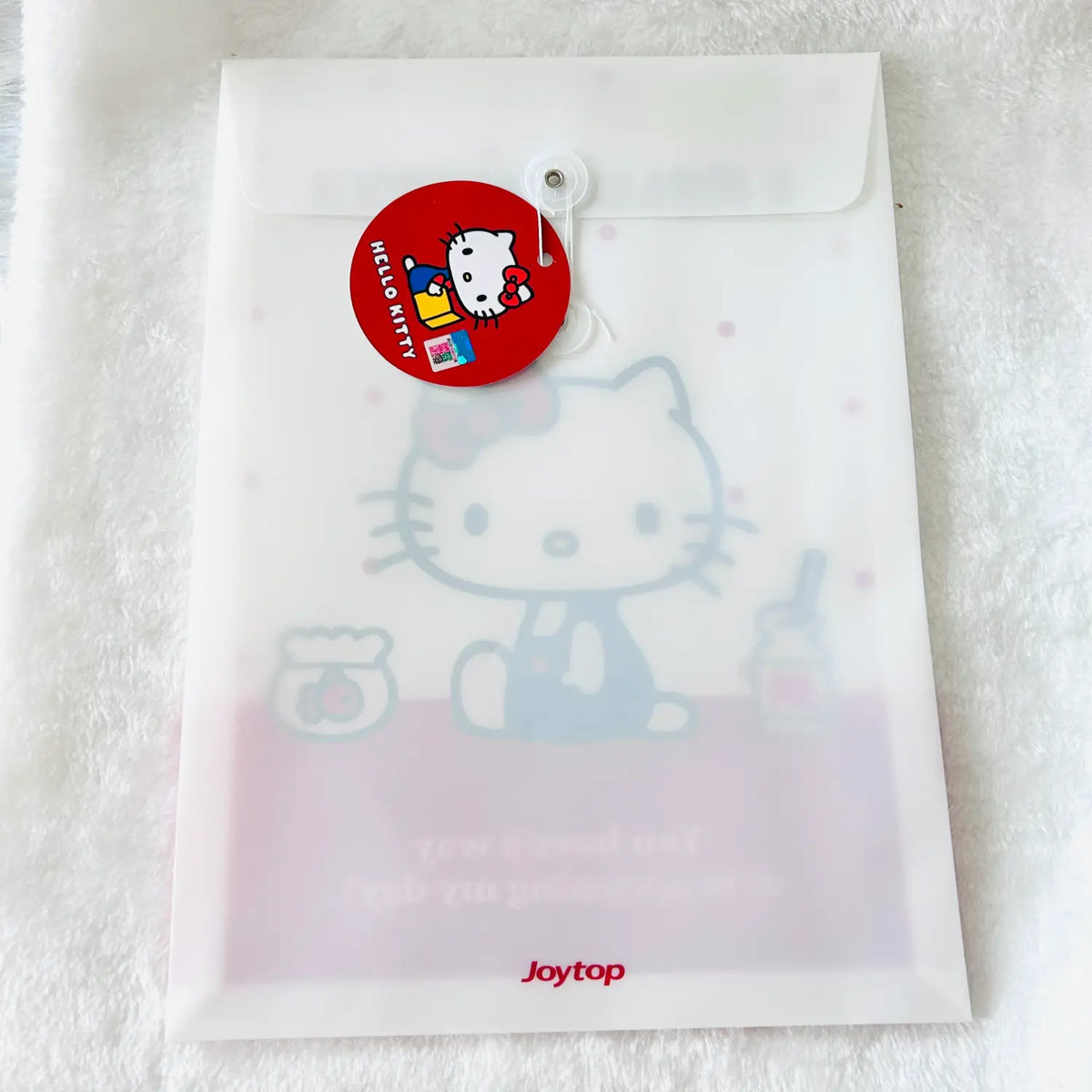 A translucent white Joytop Hello Kitty file pouch with a red round hang tag, placed on a white textured fabric background.