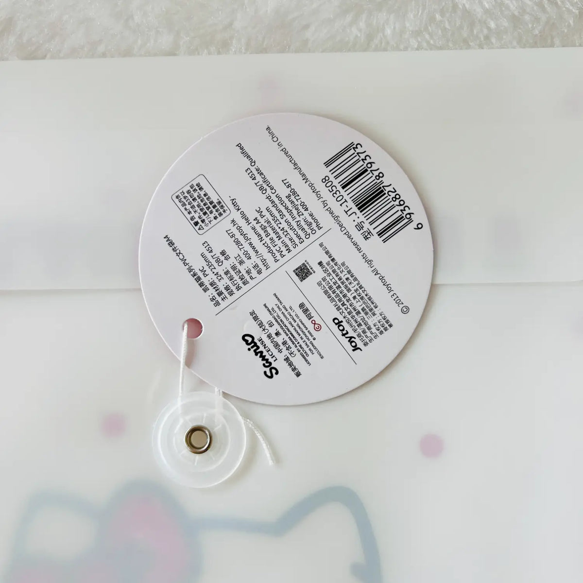 A round white product tag with a barcode and Japanese text, attached to a clear plastic loop with a gold metal ring.