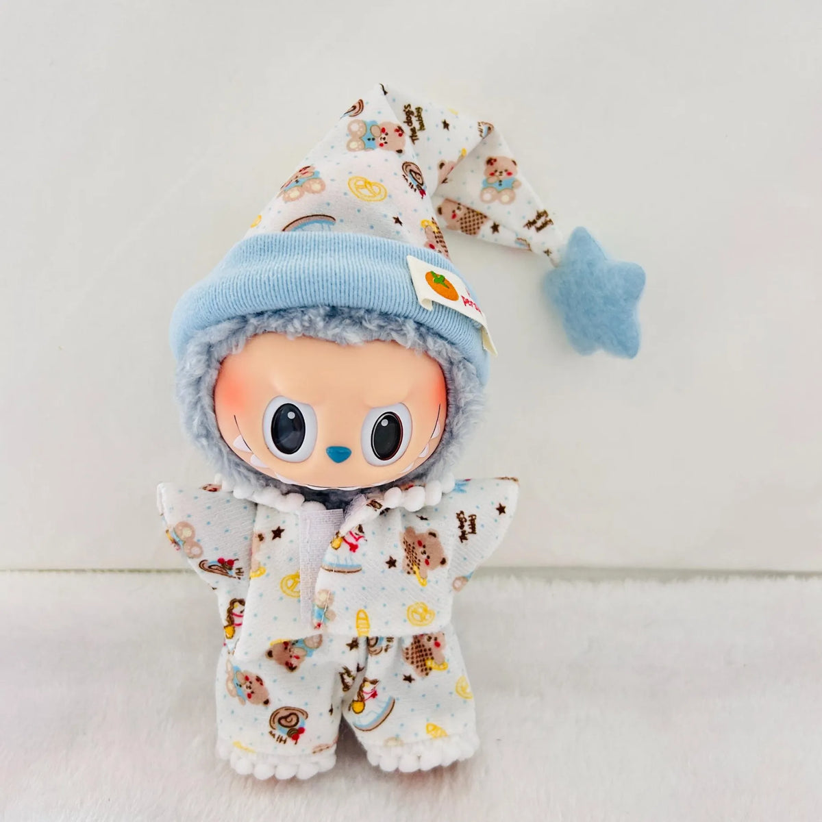 A Labubu plush doll  wearing a white pyjamas outfit with a colourful cartoon print, and a light blue knit hat.