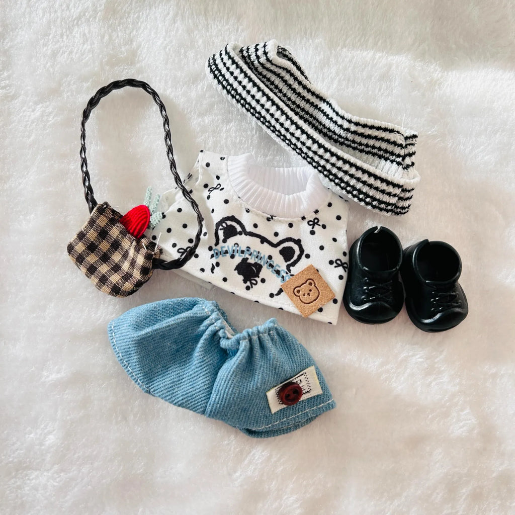 A flat-lay of a doll outfit set, including a white knit sweater with a bear face, black polka dots, a black and white striped beanie, a plaid handbag, a blue corduroy bootie, and black shoes.