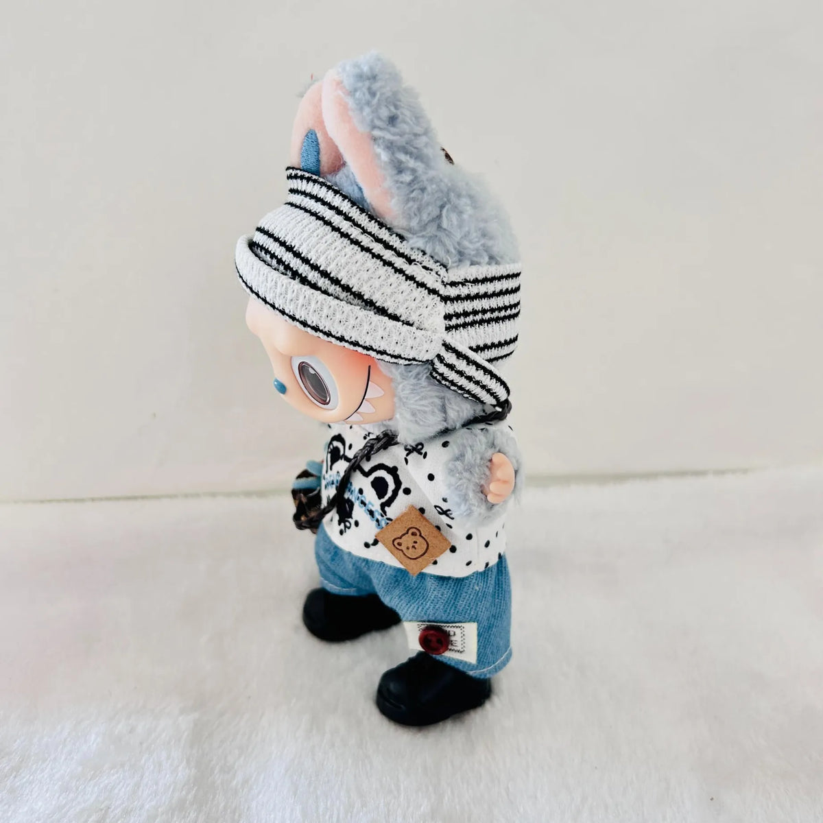 A Labubu plush doll with, a black-and-white striped knit beanie, a white patterned top, blue pants, and black shoes.