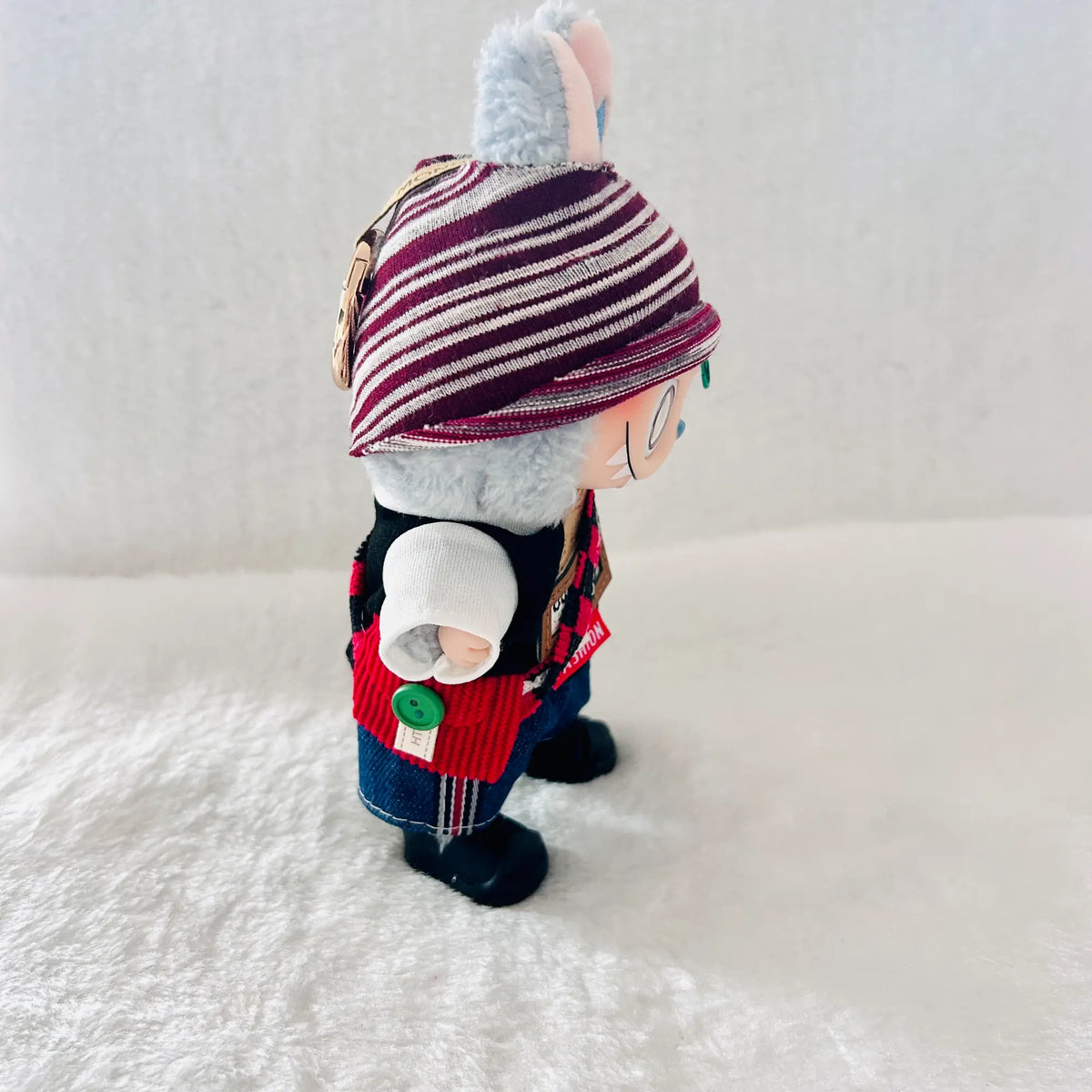 A small plush toy figure shown from the side, wearing a striped knit hat, red vest, blue pants, and black boots.