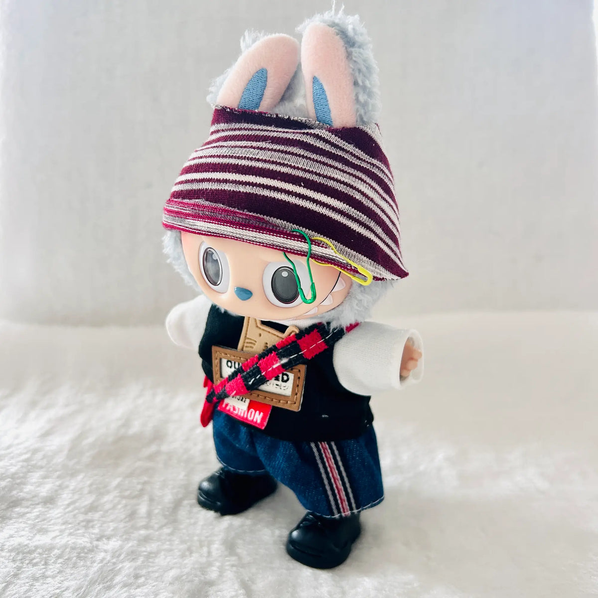 A custom handcrafted plush doll wearing a burgundy and white striped beanie, a black vest, blue jeans, and black shoes. 