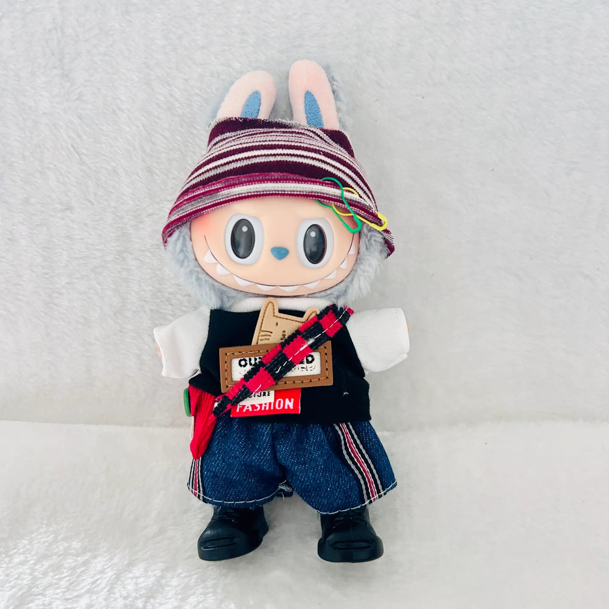 A Labubu plush doll wearing a striped knit hat, a black vest, blue denim shorts, and black shoes.