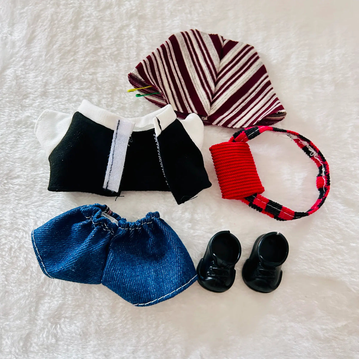 A doll outfit set including a burgundy and cream striped hat, a black top with white trim, blue denim shorts, and black shoes. A red and black patterned headband is also included.