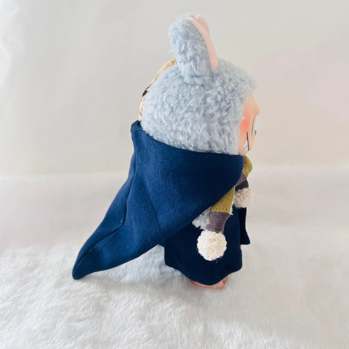 A small plush doll or character figure shown from the back, wearing a dark navy blue cape, and a yellow-green garment with white pom-poms.