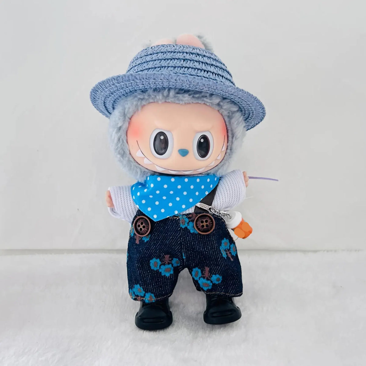 A cute doll character wearing a blue woven hat, a blue polka-dot bandana, dark blue floral overalls, and black shoes.