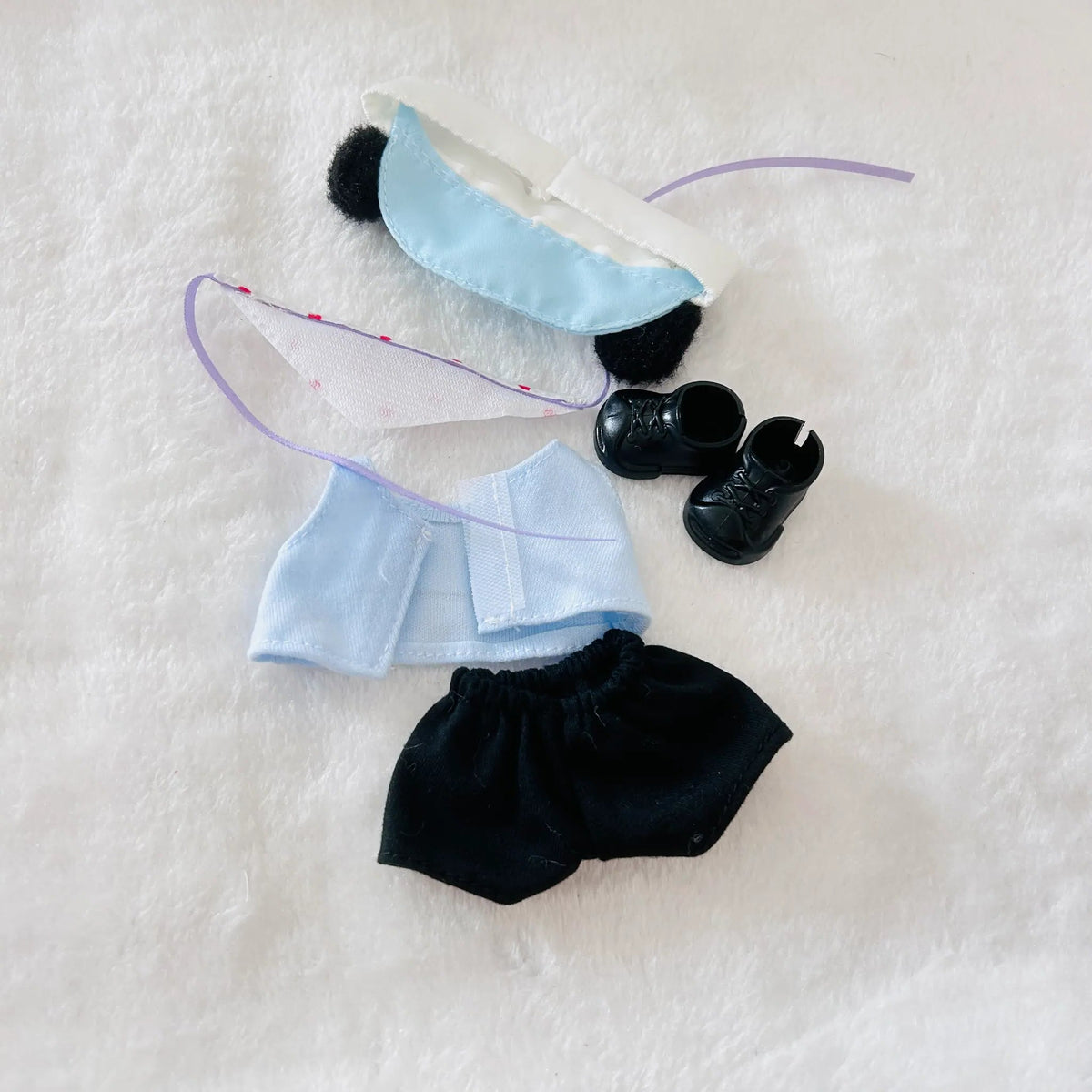 A doll outfit set laid flat on a white textured background, including a light blue and white dress, black shoes, a black bonnet, and a white skirt.