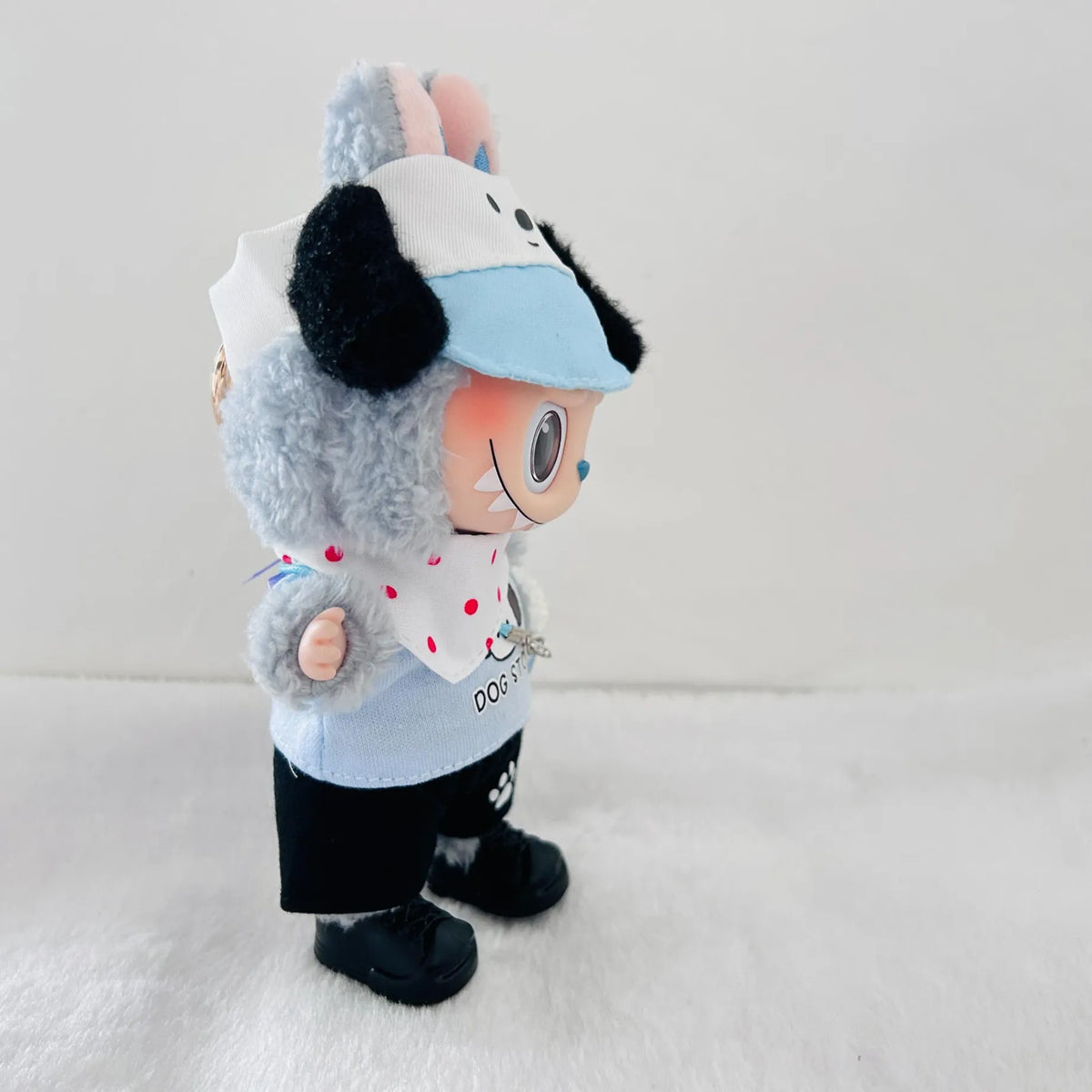 A plush doll styled like a dog character wearing a light blue and white cap, a white bib with red polka dots, a light blue shirt, black pants, and black shoes.