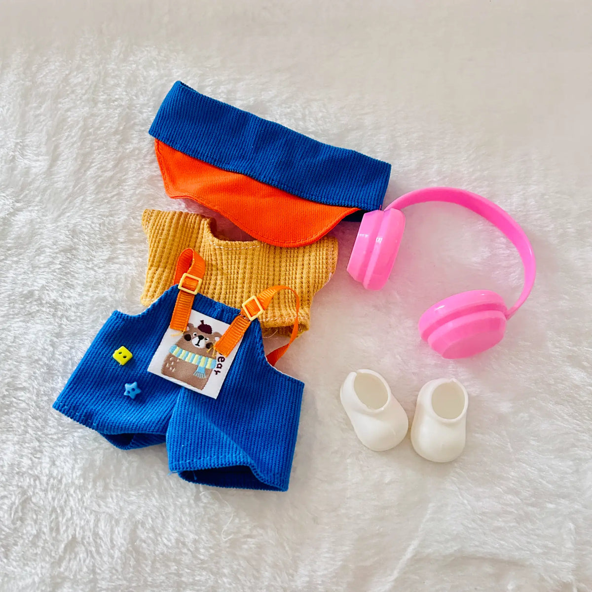 A doll clothing including a blue knit cap, orange and blue knit top, blue overalls with orange straps, pink headphones, and white booties.