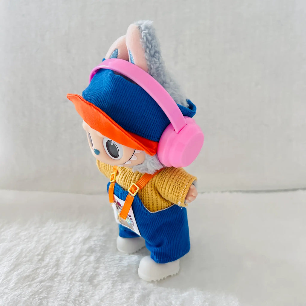 A plush toy doll dressed in a blue knit hat and overalls with a yellow sweater, orange visor, and pink headphones.
