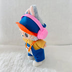 A plush toy doll dressed in a blue knit hat and overalls with a yellow sweater, orange visor, and pink headphones.
