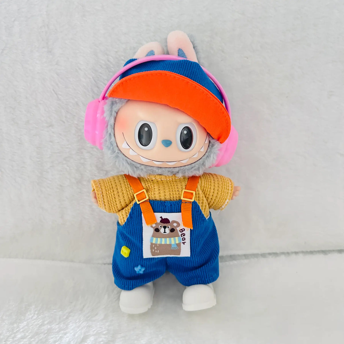 A Labubu plush toy doll dressed in a yellow knit top, blue corduroy overalls, an orange visor, and pink headphones.
