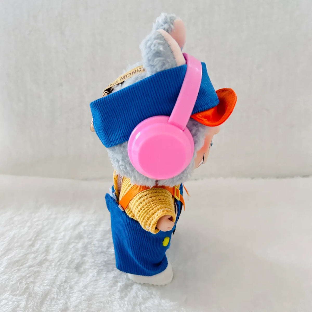 A plush toy dressed in a blue corduroy outfit with a yellow knit sweater, wearing pink over-ear headphones and an orange hat.