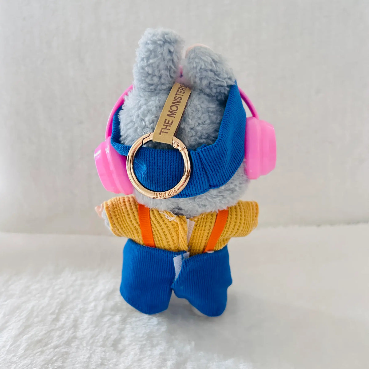 A plush toy keychain of a blue furry monster wearing pink headphones, a blue harness, a yellow sweater, and blue pants.