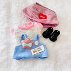 A top view of a doll outfit set, including a pink hat with a red heart patch, a pink knit top, a blue corduroy romper and small black shoes.