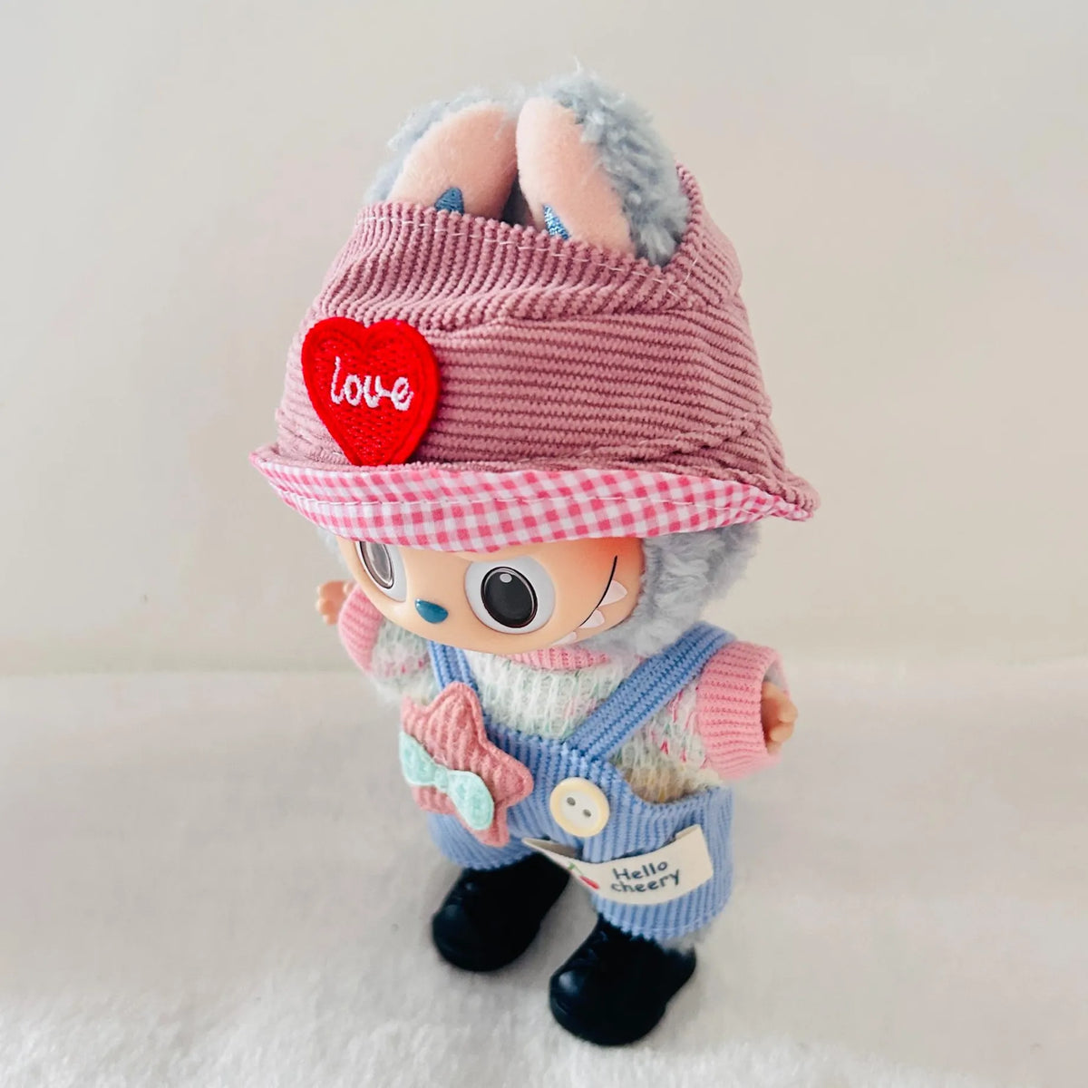 A Labubu plush doll with a pink striped hat featuring a red heart patch, dressed in pastel pink and blue overalls with black shoes.