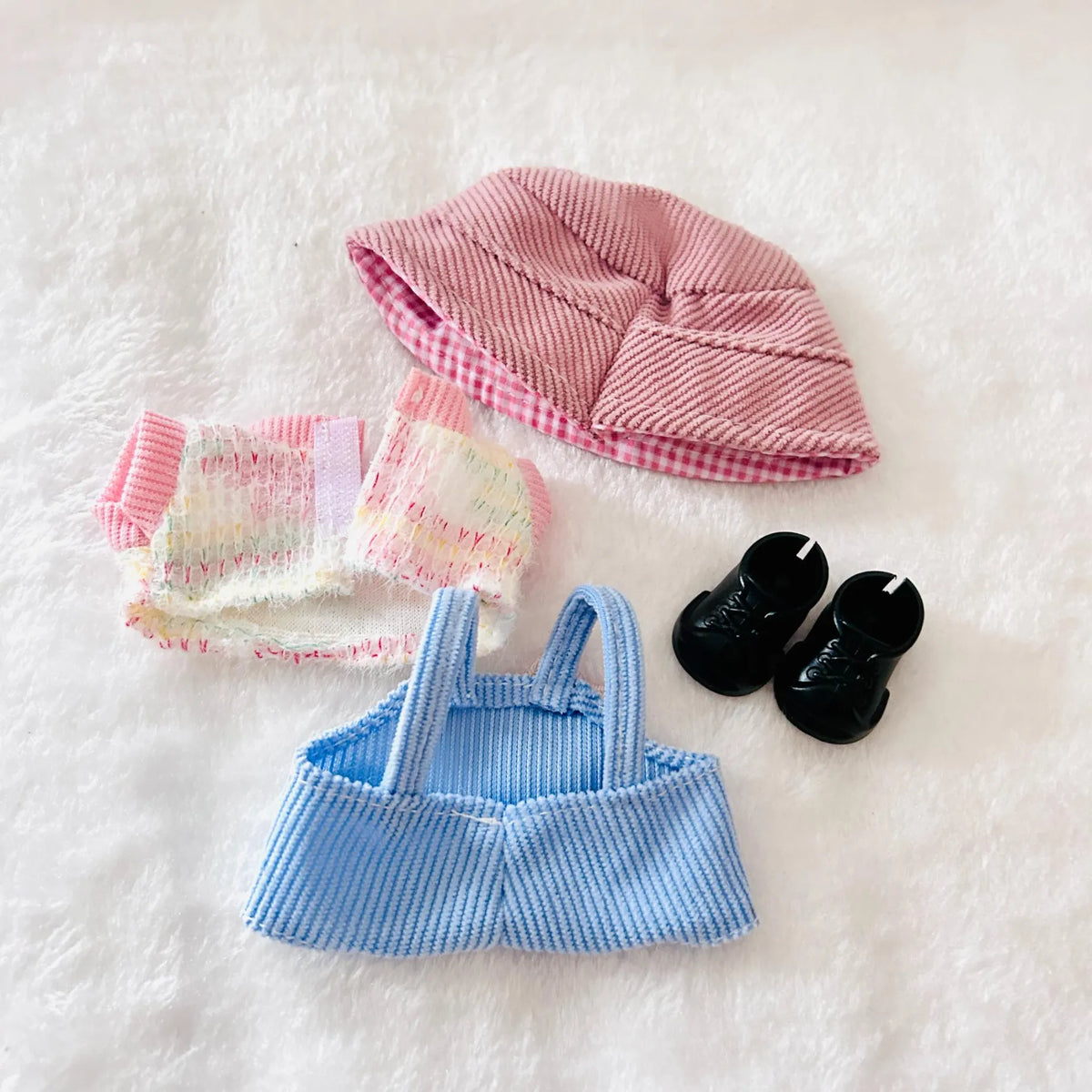A back view of a doll clothing set, including a pink hat, a pastel striped pants, a light blue striped top, and black shoes.