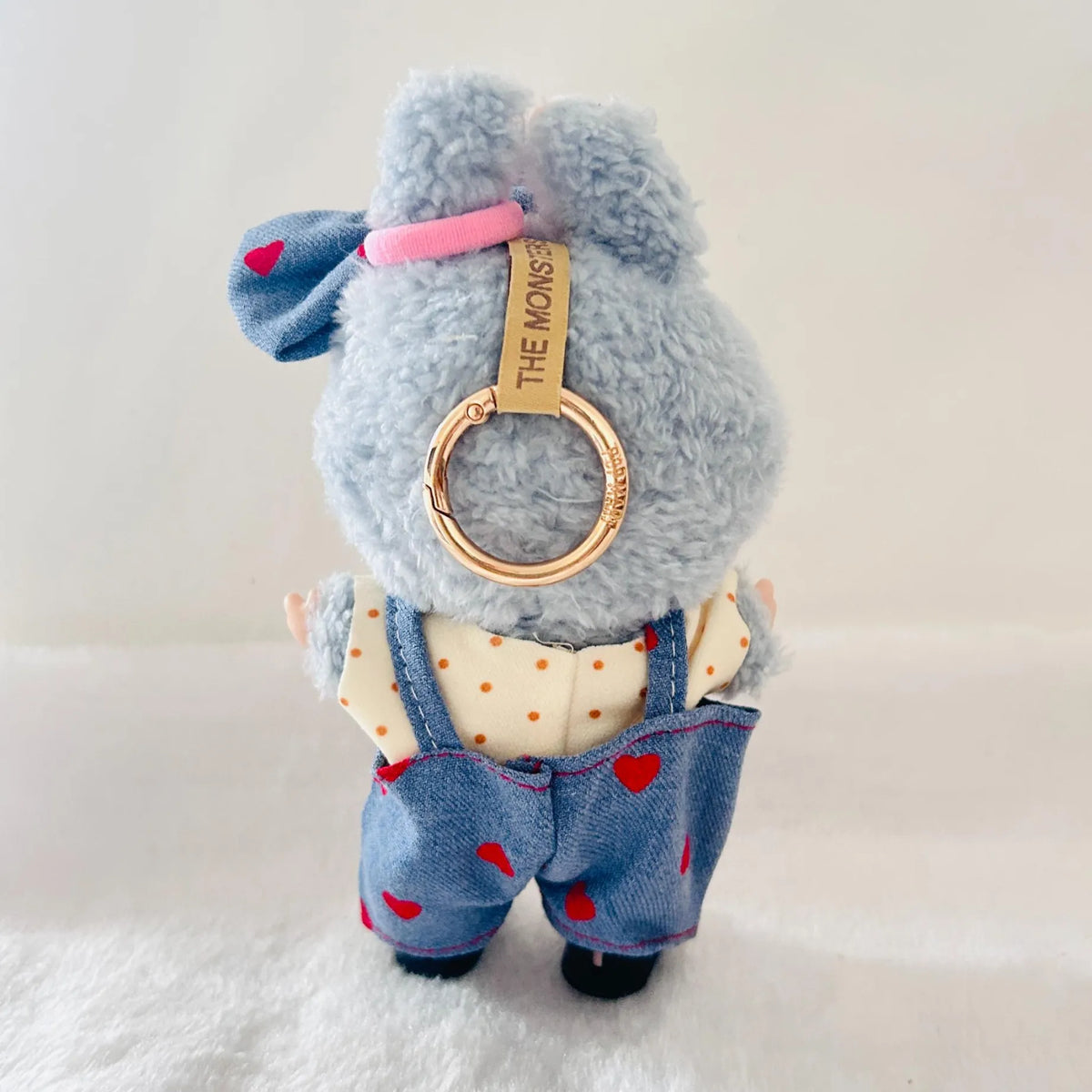 A back view of a Labubu plush doll wearing a denim overalls with red hearts, a cream polka-dot shirt.