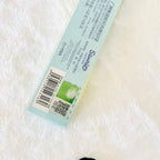 A close-up of a Scowl brand product box lying on a white textured fabric background. The box is light blue with black text, a barcode, a QR code, and a small green square graphic.
