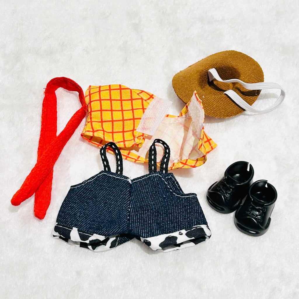 A flat-lay of doll clothing including a tan hat with a white strap, a pair of black doll boots, a yellow-and-orange plaid top, a denim-and-white cow-print overalls.