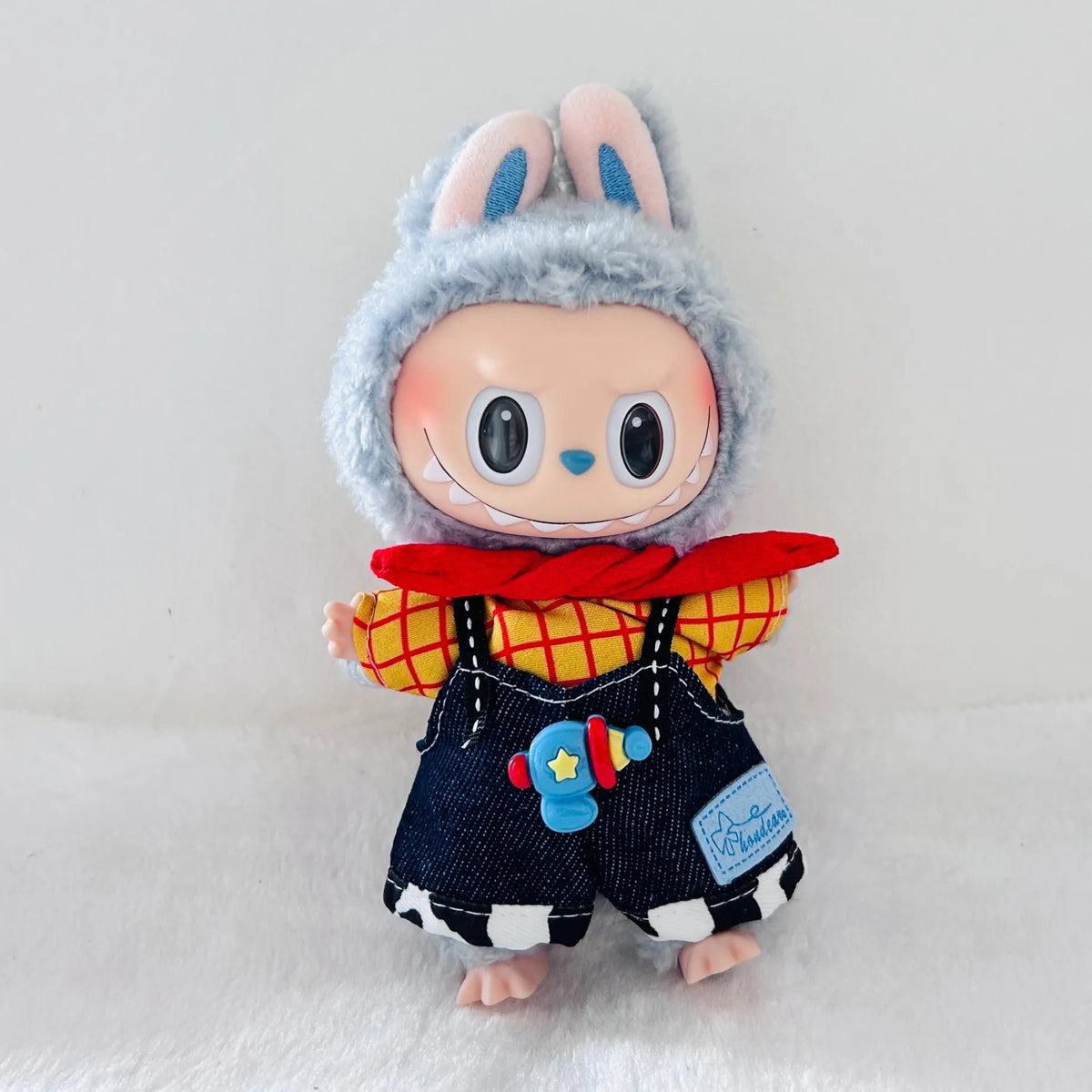 A Labubu plush doll wearing a red and yellow plaid shirt, dark blue denim-style overalls with a blue toy blaster patch.
