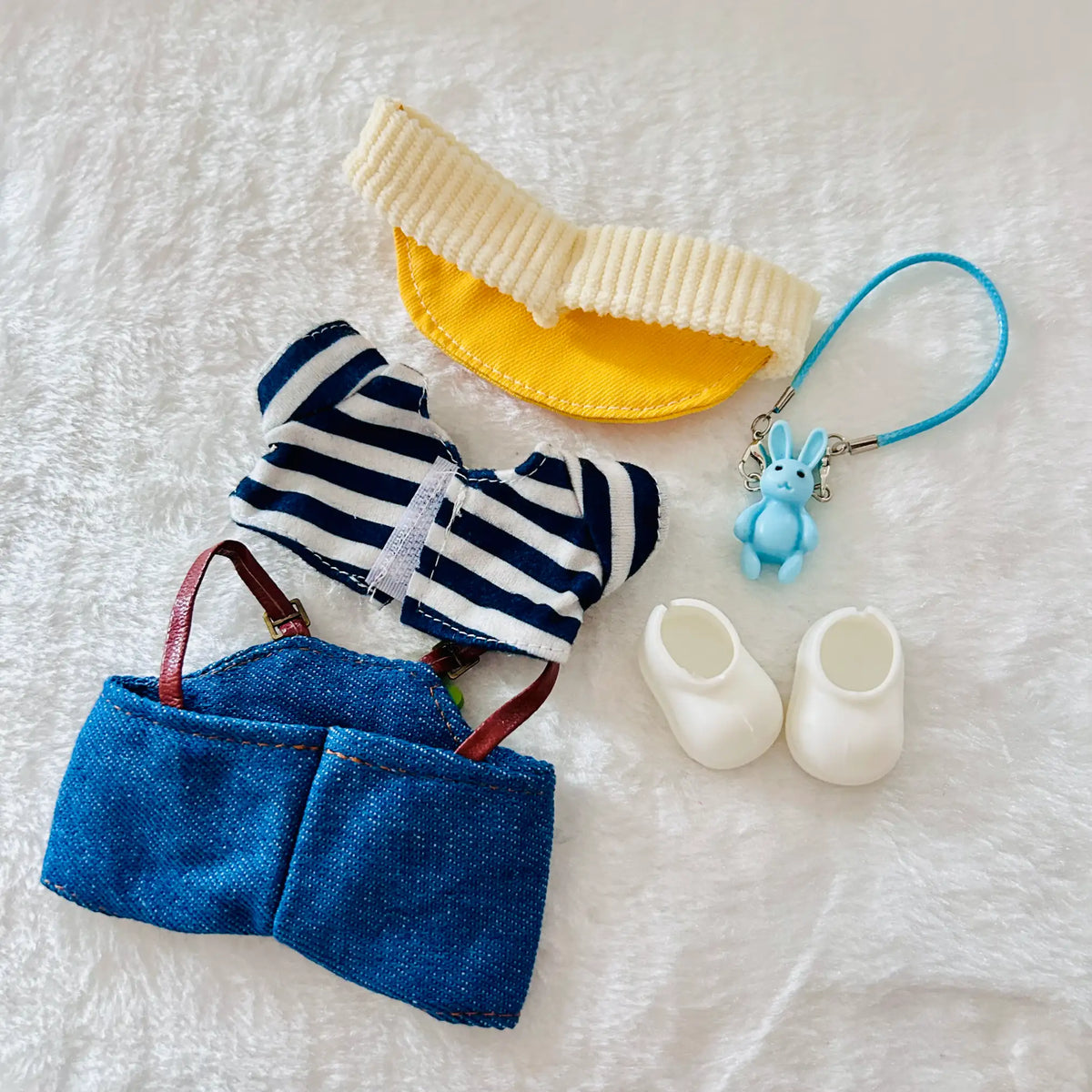 A doll or miniature clothing set including a cream ribbed bonnet, a navy-and-white striped top, blue denim shorts with brown straps, a blue bunny charm keychain, and a pair of small white booties.