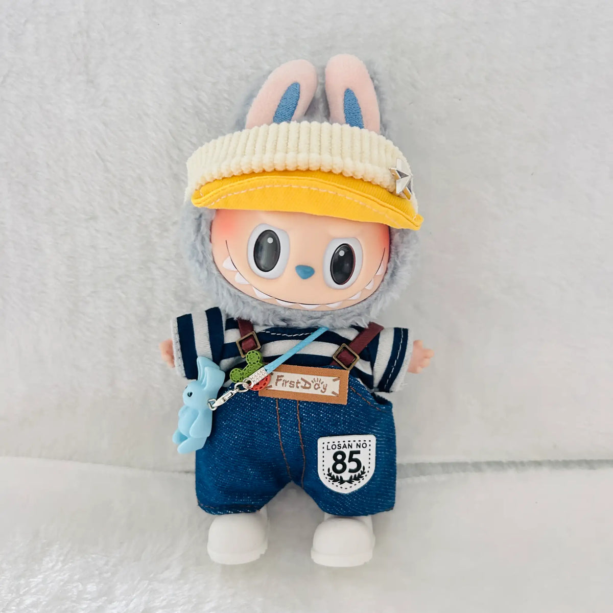 A Labubu plush doll wearing a cream ribbed cap, blue denim-style overalls with brown suspenders, a navy-and-white striped shirt, and white shoes.