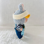 A plush rabbit doll wearing a cream ribbed knit beanie with a yellow ear, a navy striped shirt, and blue overalls.