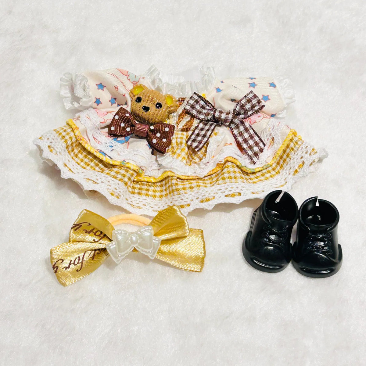 A Labubu doll outfit set featuring a cream and yellow gingham skirt with lace trim, a small teddy bear accessory, a bow and a pair of black doll shoes.