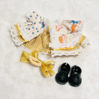 A back view of a Labubu doll dress laid with  with blue stars and orange animals, a yellow satin bow, and a pair of small black shoes.