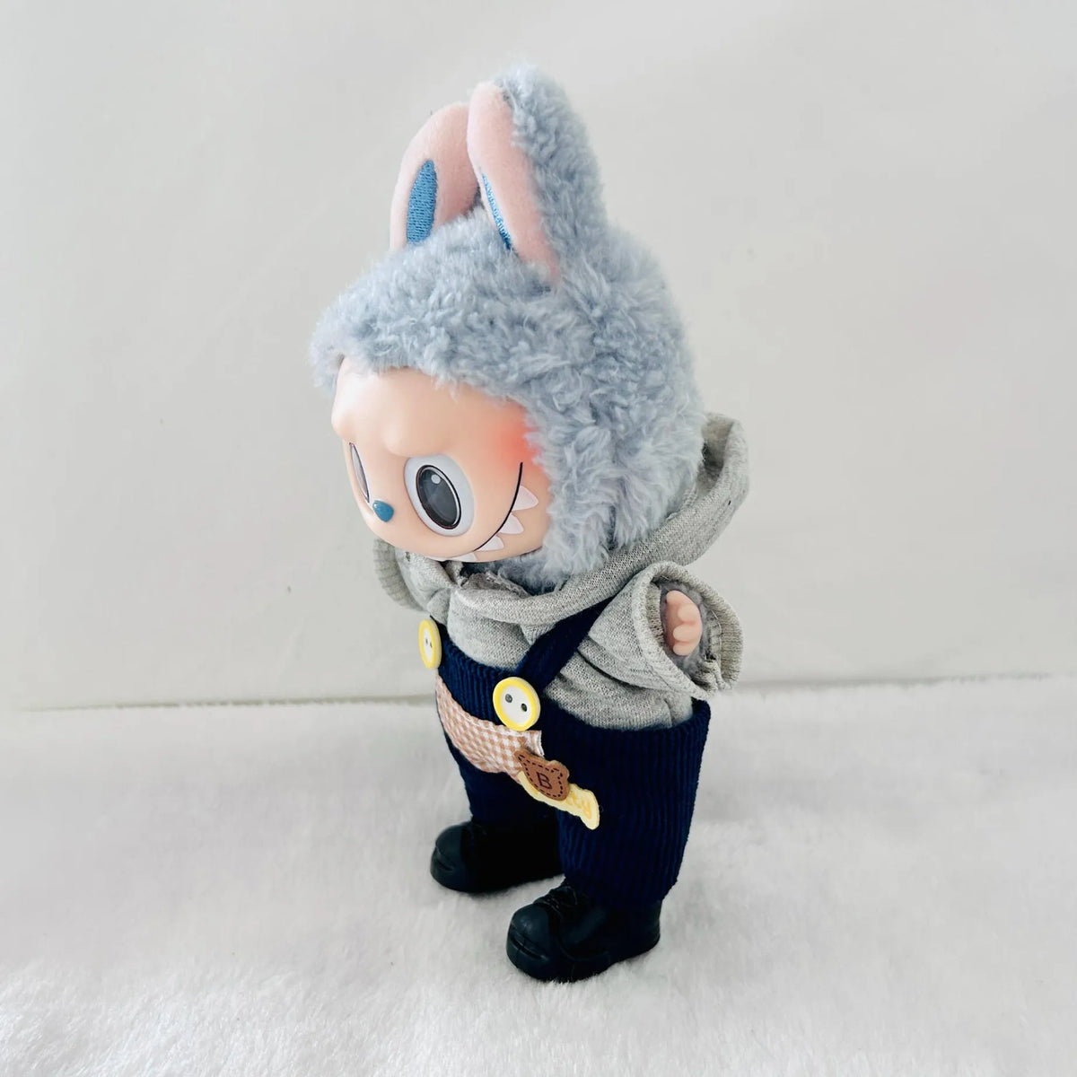 A Labubu plush doll  wearing a grey hoodie, and dark blue overalls, standing on a white textured surface.