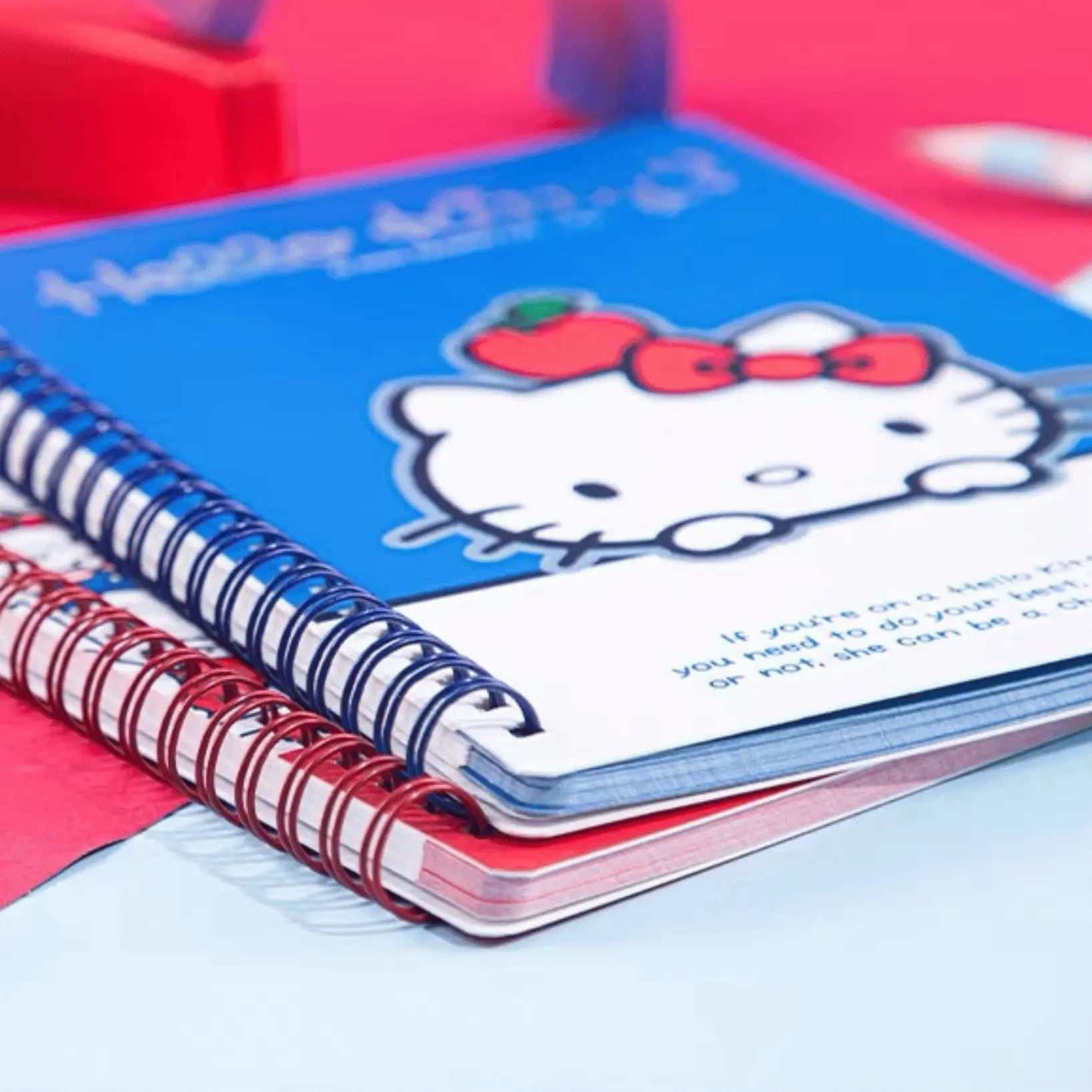 A Hello Kitty spiral notebook with a blue cover and white pages, stacked on top of another red spiral notebook.