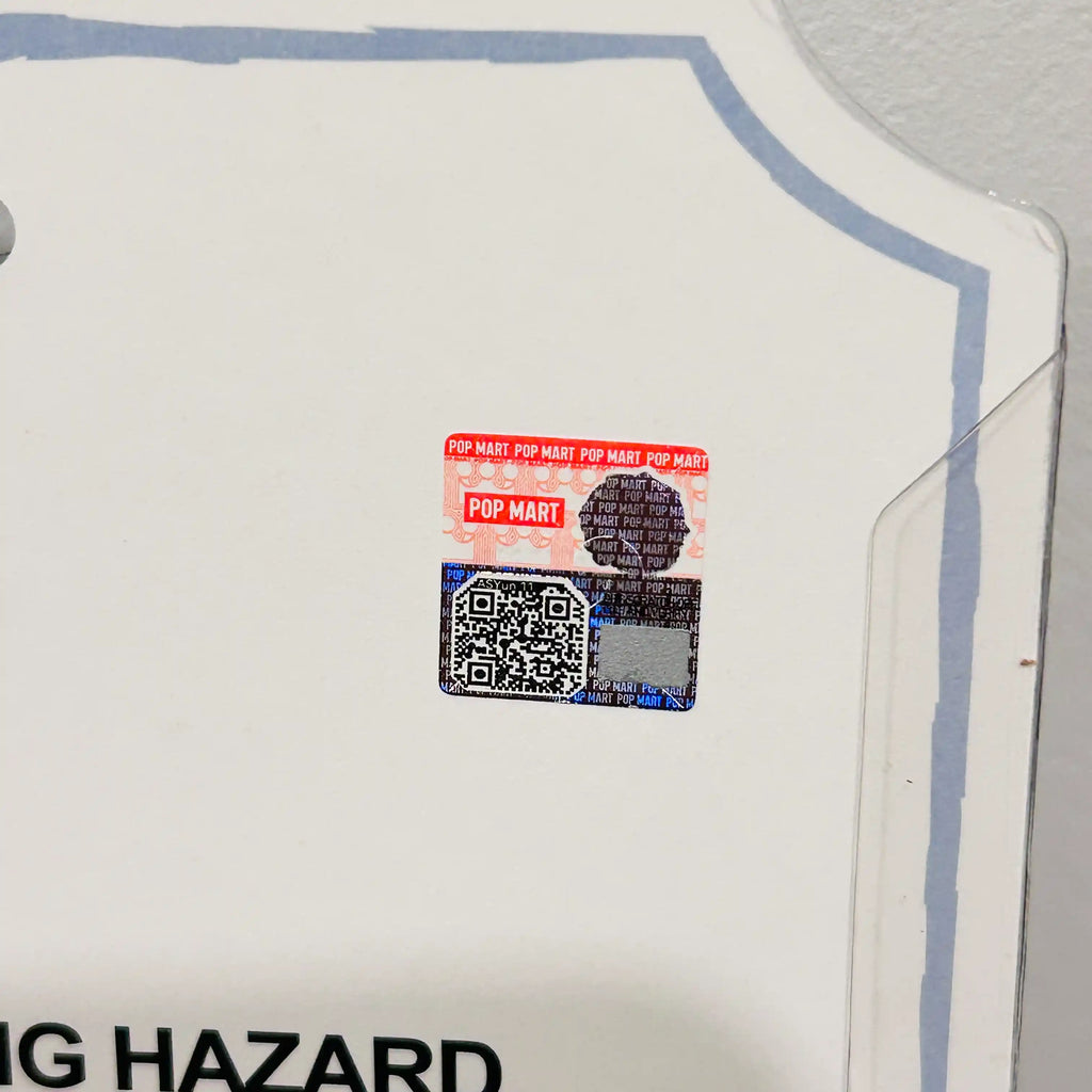A close-up of a white plastic retail display card with a blue border, showing a small Pop Mart holographic security sticker near the center.