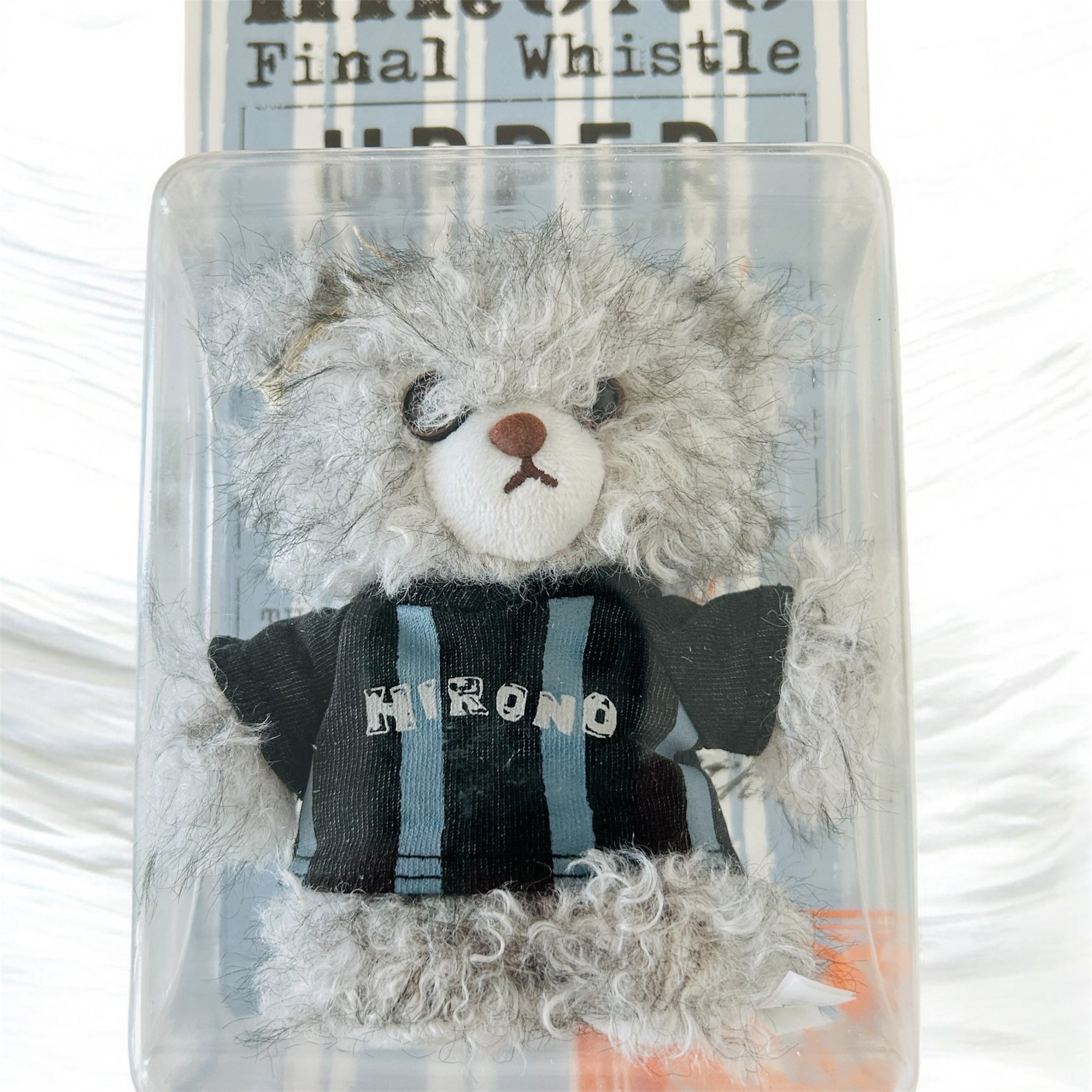A small gray plush teddy bear with a white snout and black embroidered eyes, wearing a black and blue striped shirt with the word 'HIRONO' on the front, displayed inside a clear plastic box.