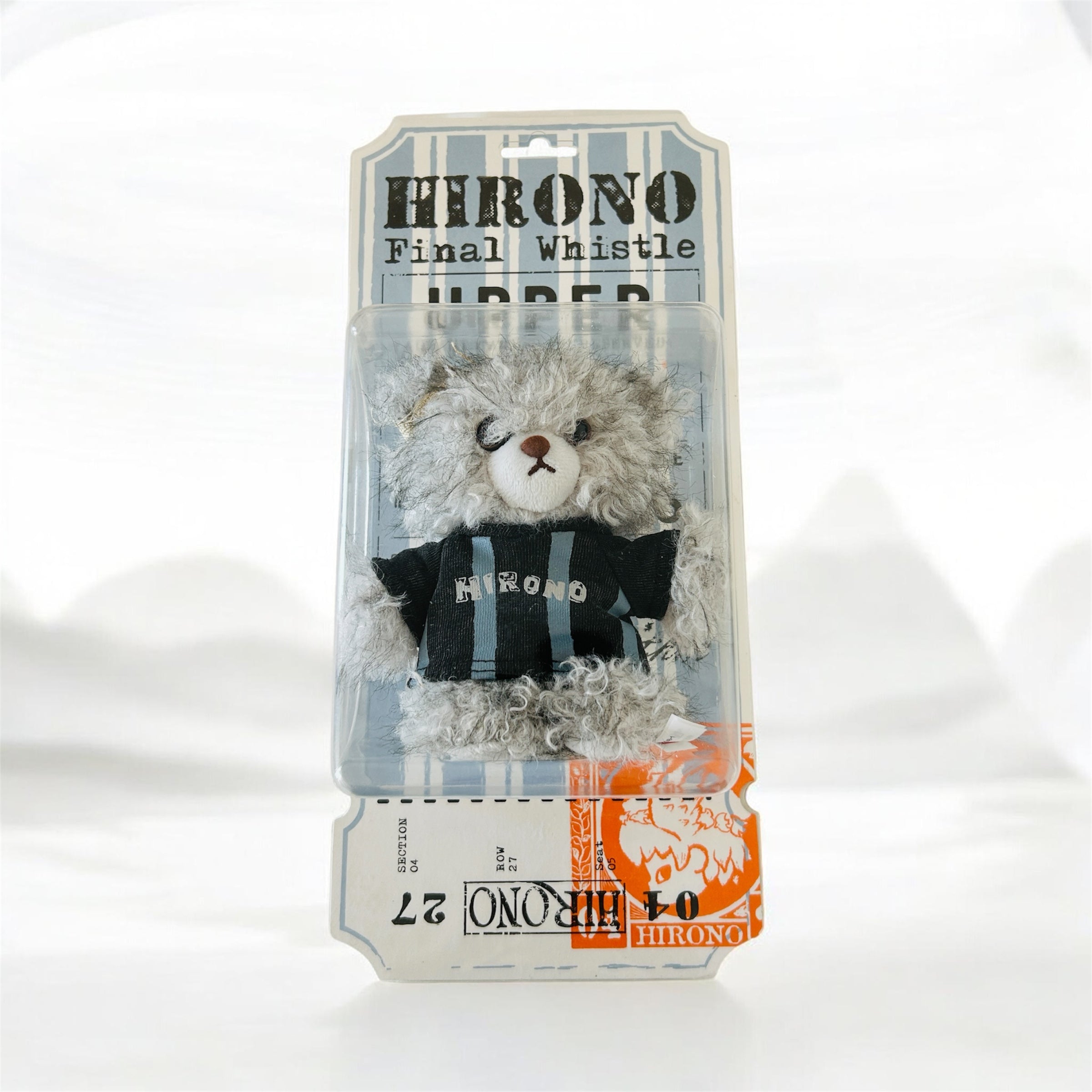 A packaged plush toy resembling a gray furry character wearing a black shirt with blue stripes and the word 'HIRONO' on the front, mounted on a printed backing card.