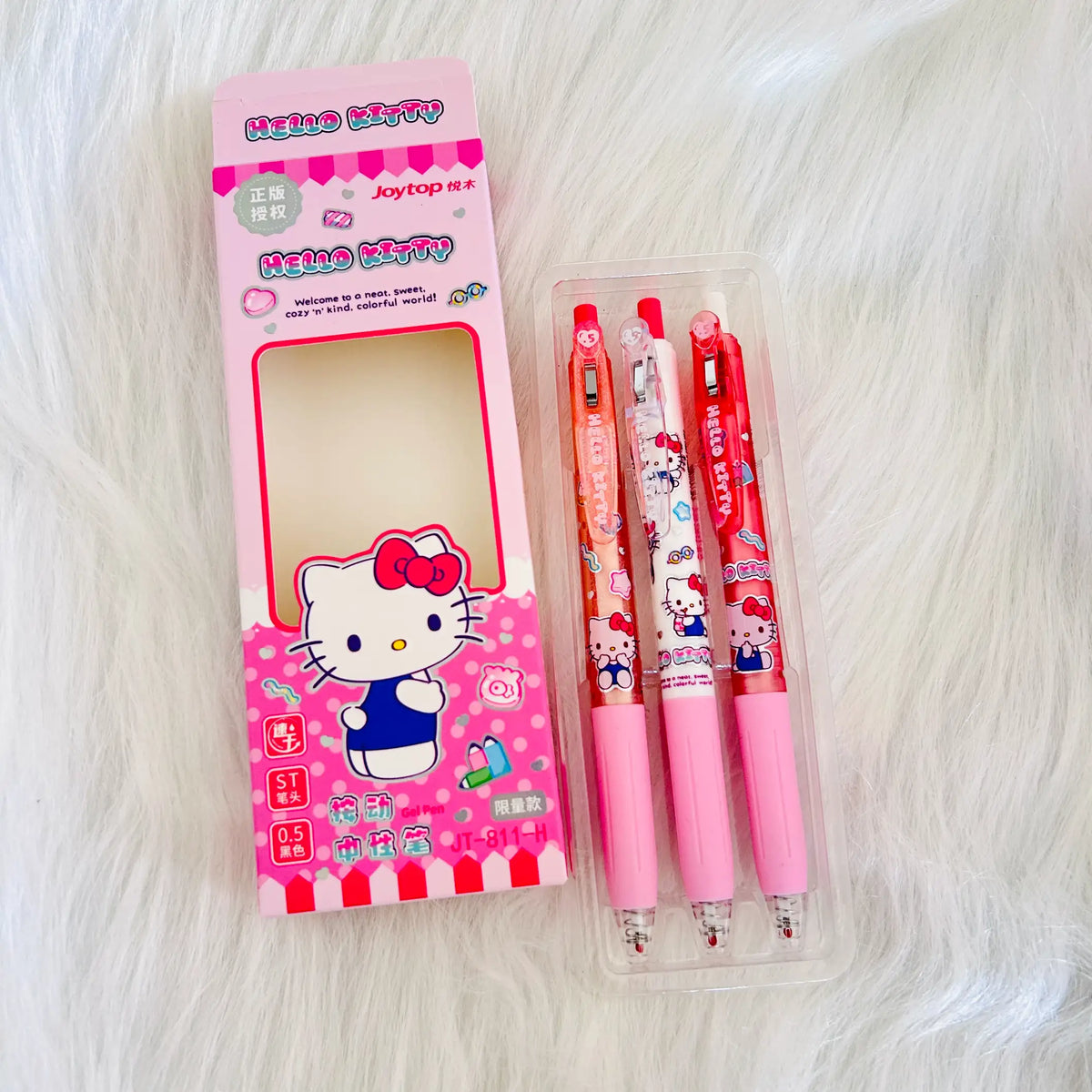 Hello Kitty themed stationery set featuring a pink branded box and three cartoon-print retractable pens in a clear plastic case, placed on a white faux-fur surface.