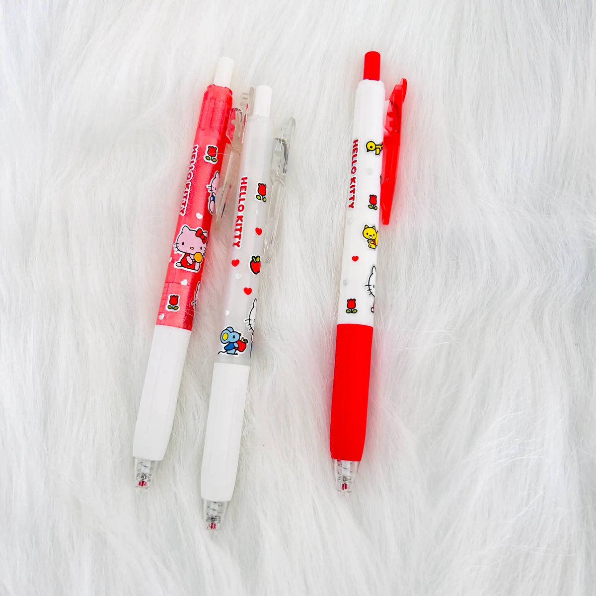 Three Hello Kitty gel pens with white and red colors and character illustrations, arranged on a white faux fur background.