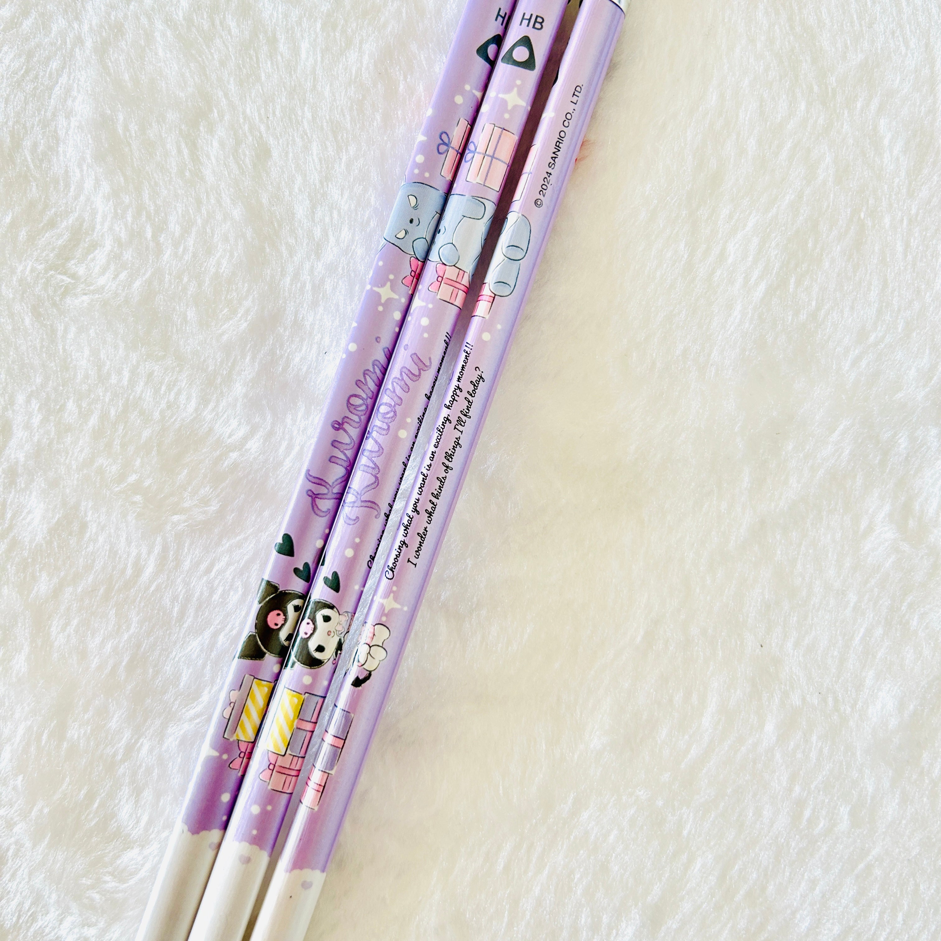 Three pastel purple stationery pencils with Sanrio character illustrations, including Kuromi, placed diagonally on a white textured faux fur surface.