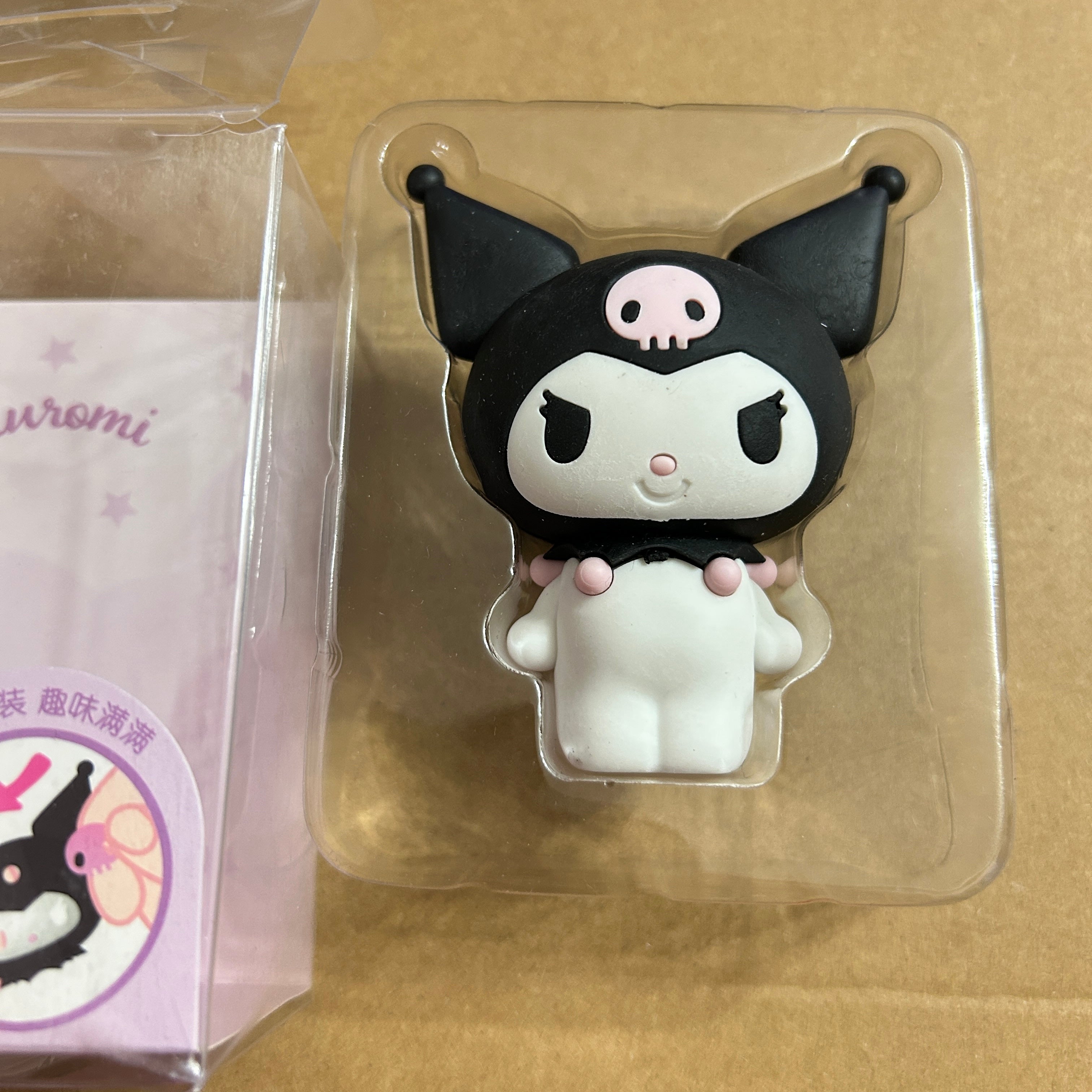 Kuromi character novelty item in clear plastic packaging, shown next to its branded box on a tan surface.